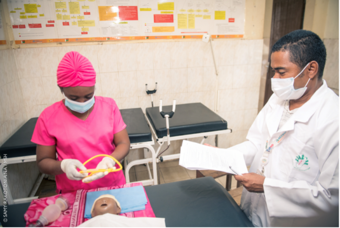 Una enfermera con una bata rosa practica habilidades en una muñeca NeoNatalie, supervisada por un técnico con una bata blanca de médico.