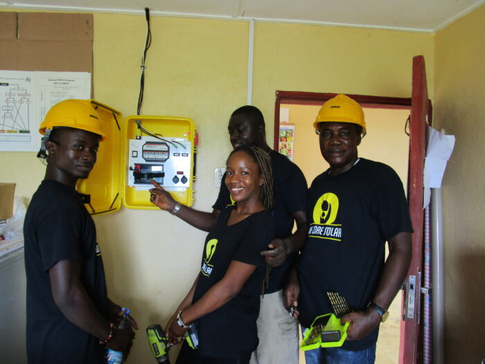 Four We Care Solar technicians checking on an installed Solar Suitcase inside a health facility. They are all looking at the camera and smiling, wearing yellow hard hats.