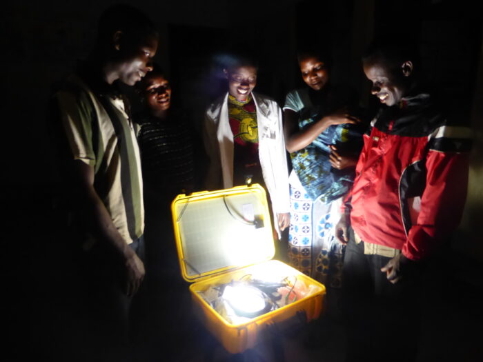 A group of Tanzanian health care workers standing around a yellow box containing a "solar suitcase" which provides solar power to their health facility. They are standing in darkness with the only light coming from an LED in the suitcase.